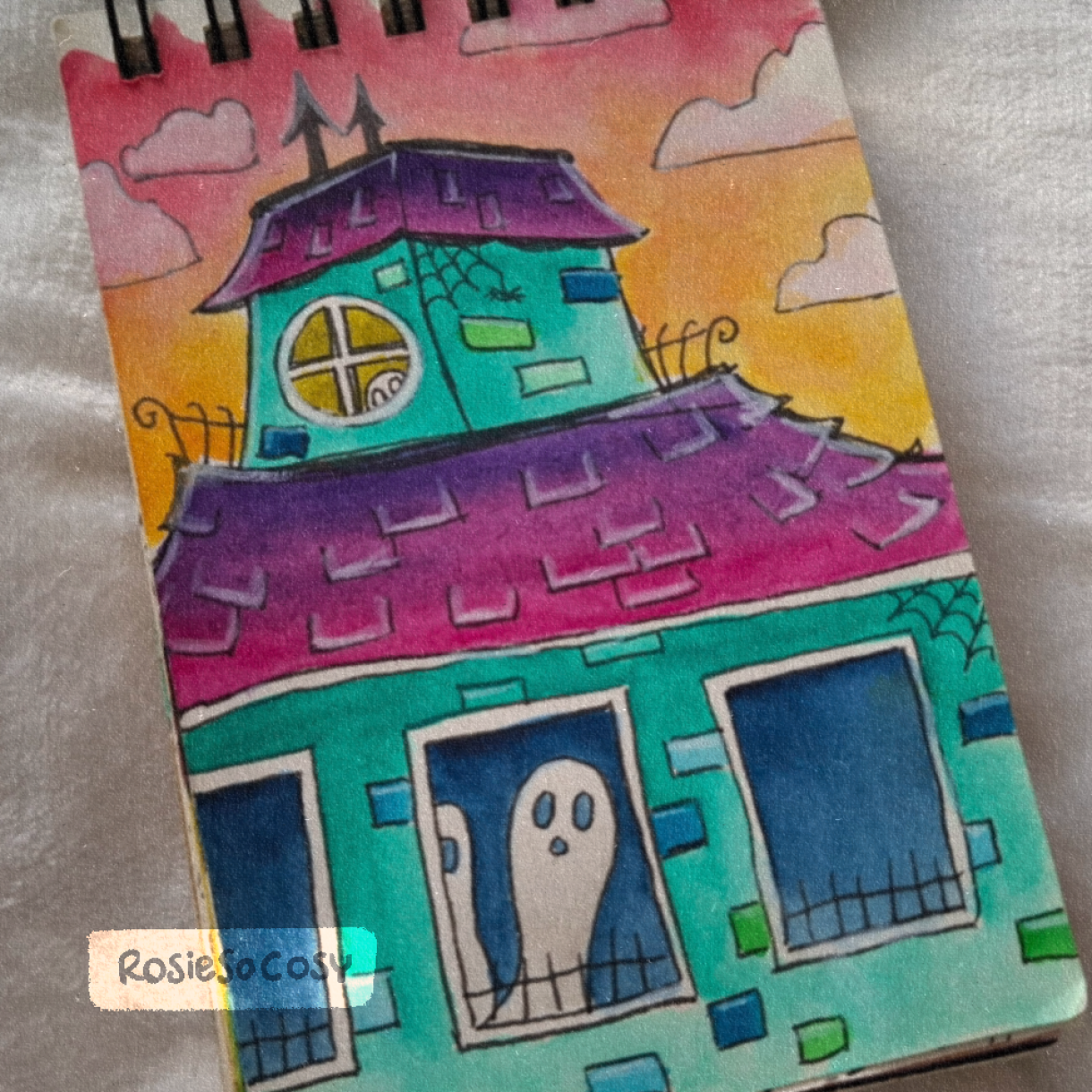 Watercolour illustration of a turquoise haunted house with pinkish purple rooftops, black gothic architectural details, and white ghosts roaming inside the building. The sky is pink and orange, indicating a sunset.